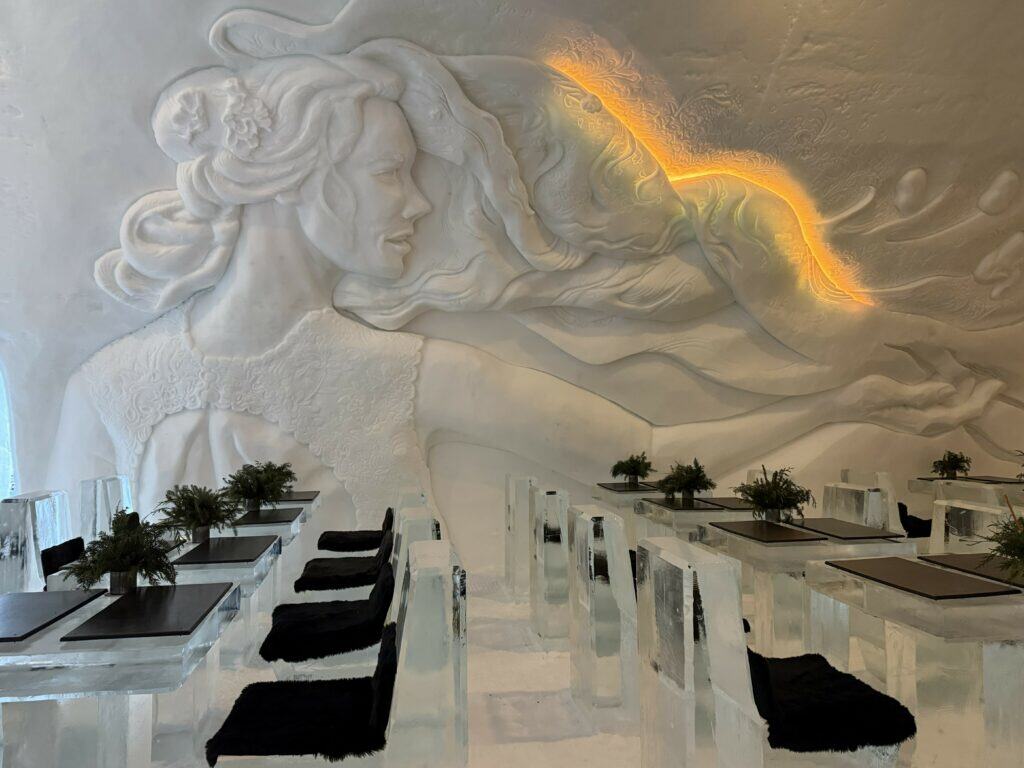 restaurant at quebec ice hotel with image of woman sculpted into back wall, tables and chairs made of ice with dark fur seatpads and table set with dark greenery and placemats.