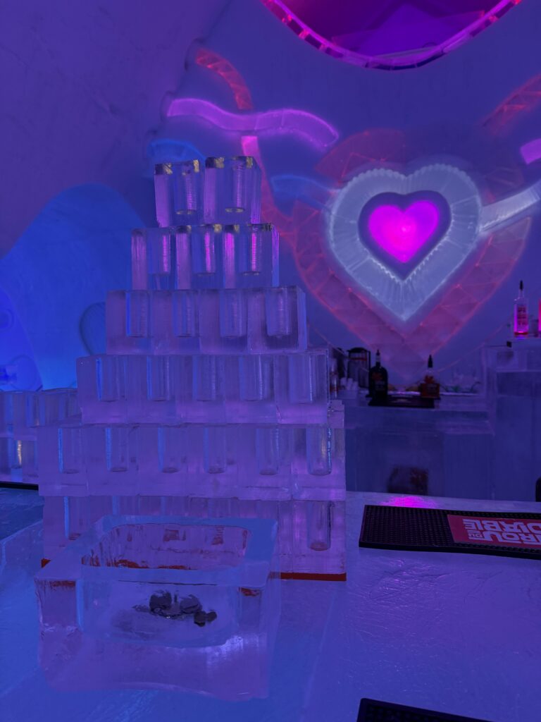 tower of ice glasses stacked on ice bar with bottles of alcohol in background - pink and purple heart on wall.