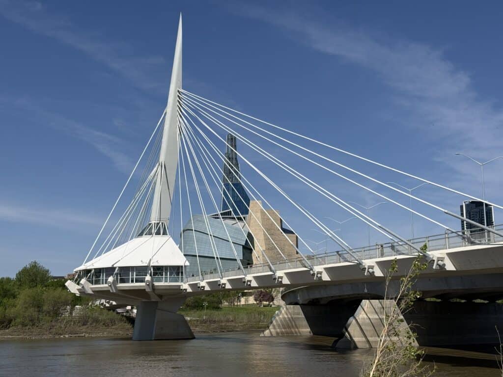 Esplanade Riel pedestrian bridge with Canadian Museum for Human Rights in background - white cables and structure set against a bright blue sky with wispy white clouds.