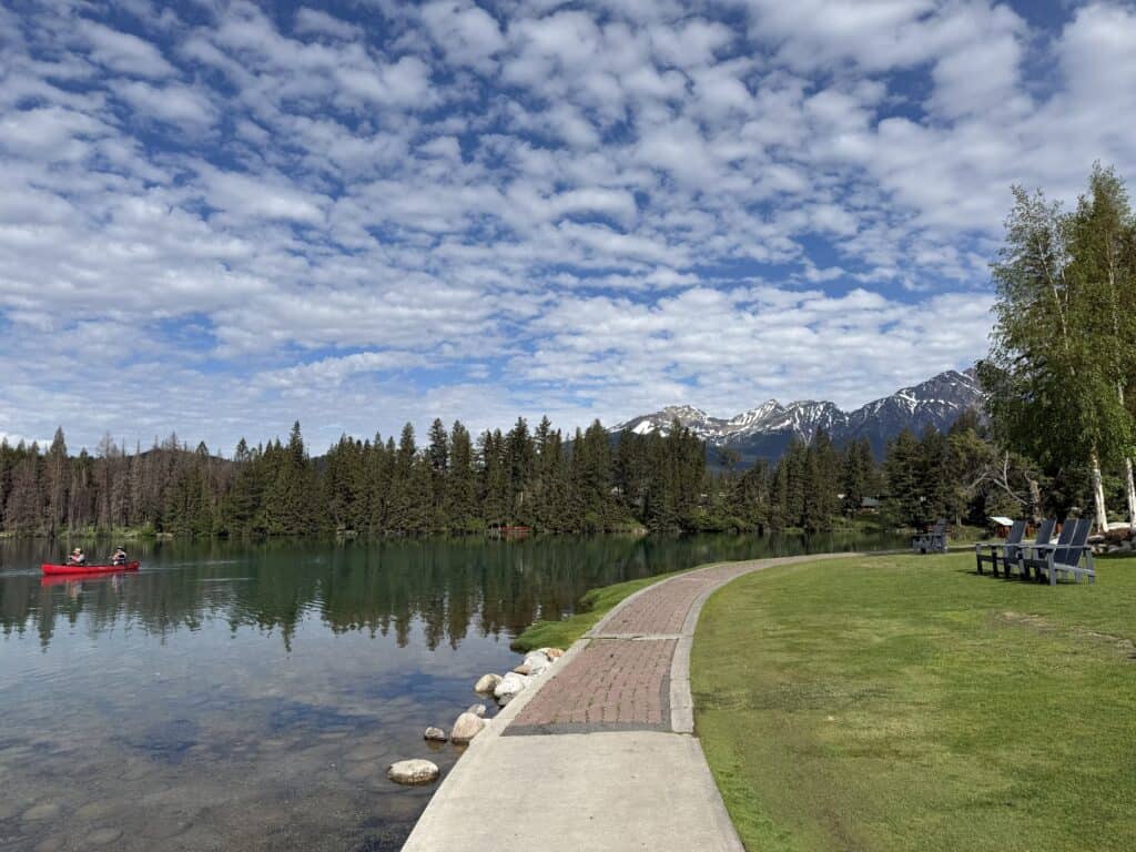 lakefront at Jasper Park Lodge in morning with evergreen trees reflecred in water and red canoe on the water - blue sky with white fluffy clouds.