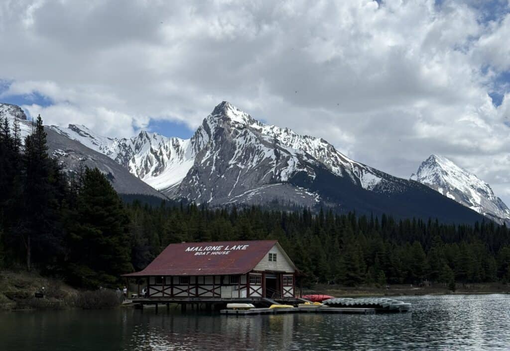 Maligne Lake Boat House at midday with snow-capped mountains and evergreen in background and several canoes by dock.