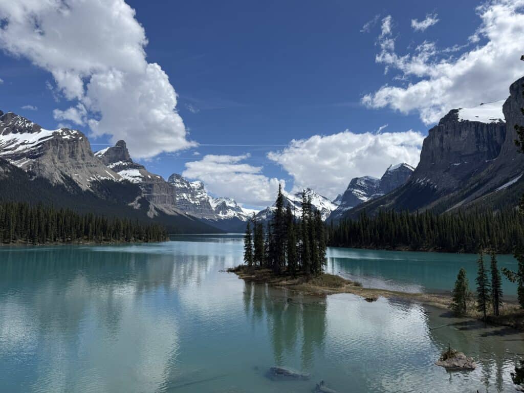 small island with evergreen trees in lake with blue/green water with snow-capped mountains in the background and blue sky with large white fluffy clouds.