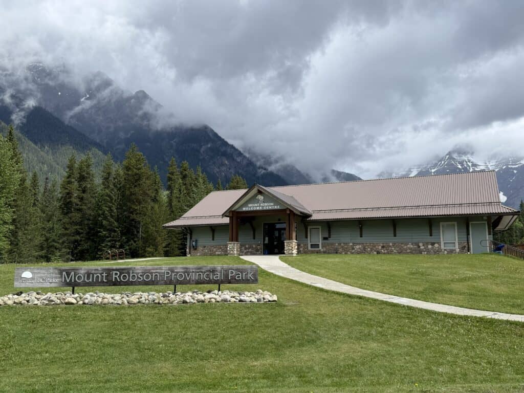 Mount Robson Provincial Park sign in front of Welcome Centre with snow-capped mountains in background and low-hanging grey clouds on an overcast day.