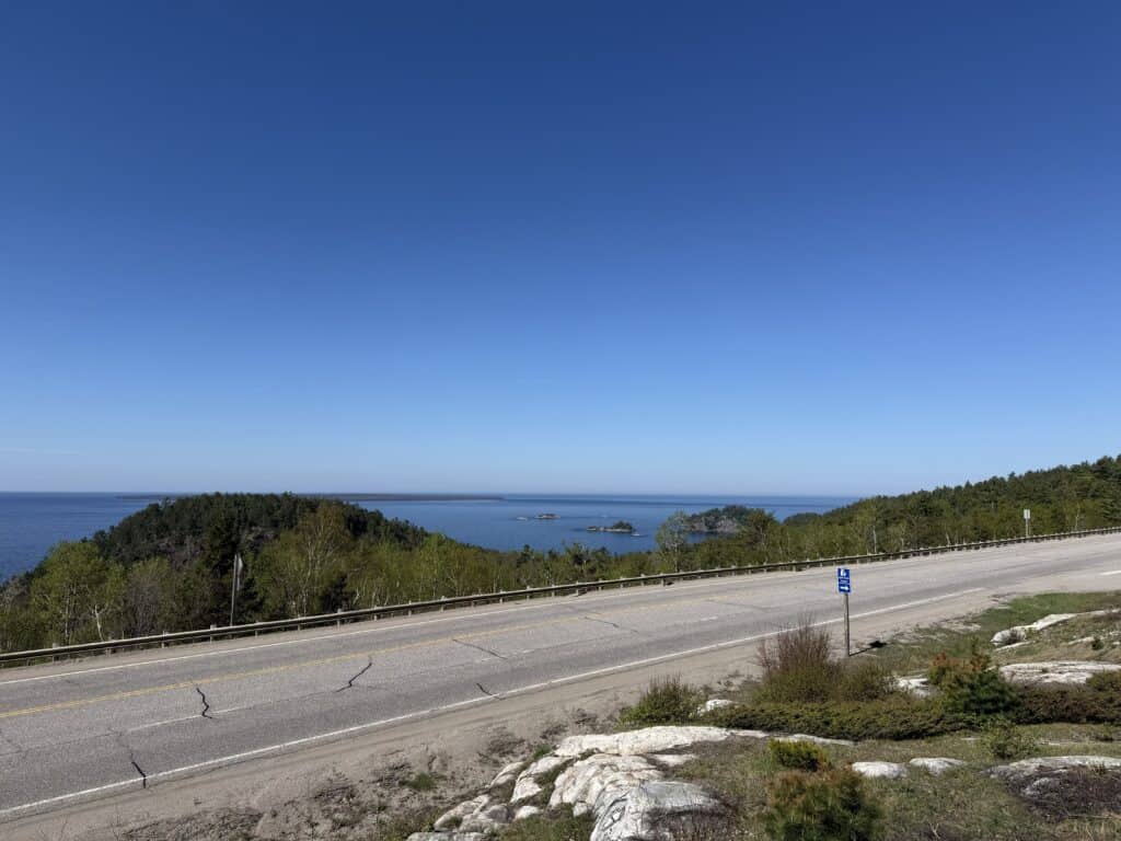 stretch of highway with rocky Canadian shield on one side and trees and lake on the other with a clear blue sky.