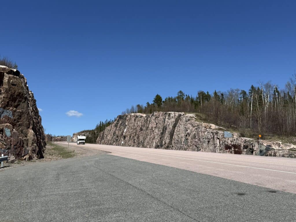 white camper driving on highway between two large rock cuts with bright blue sky.