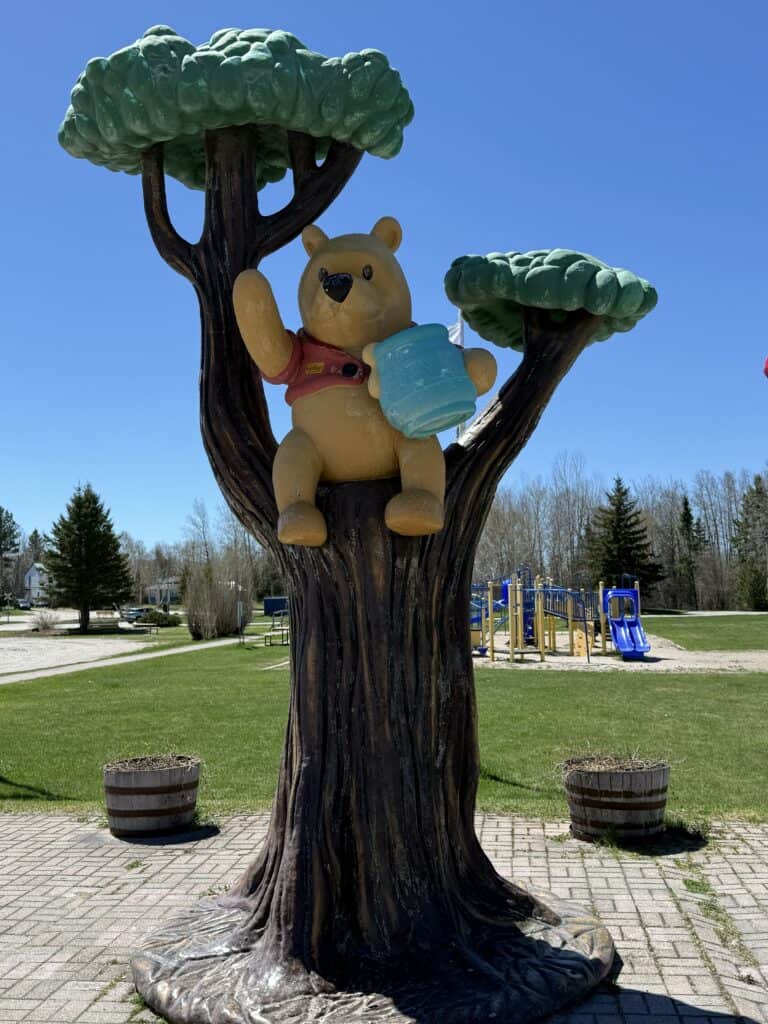 large sculpture of Winnie-the-Pooh sitting in tree holding blue honey pot on a day with bright blue sky with playground visible in background.