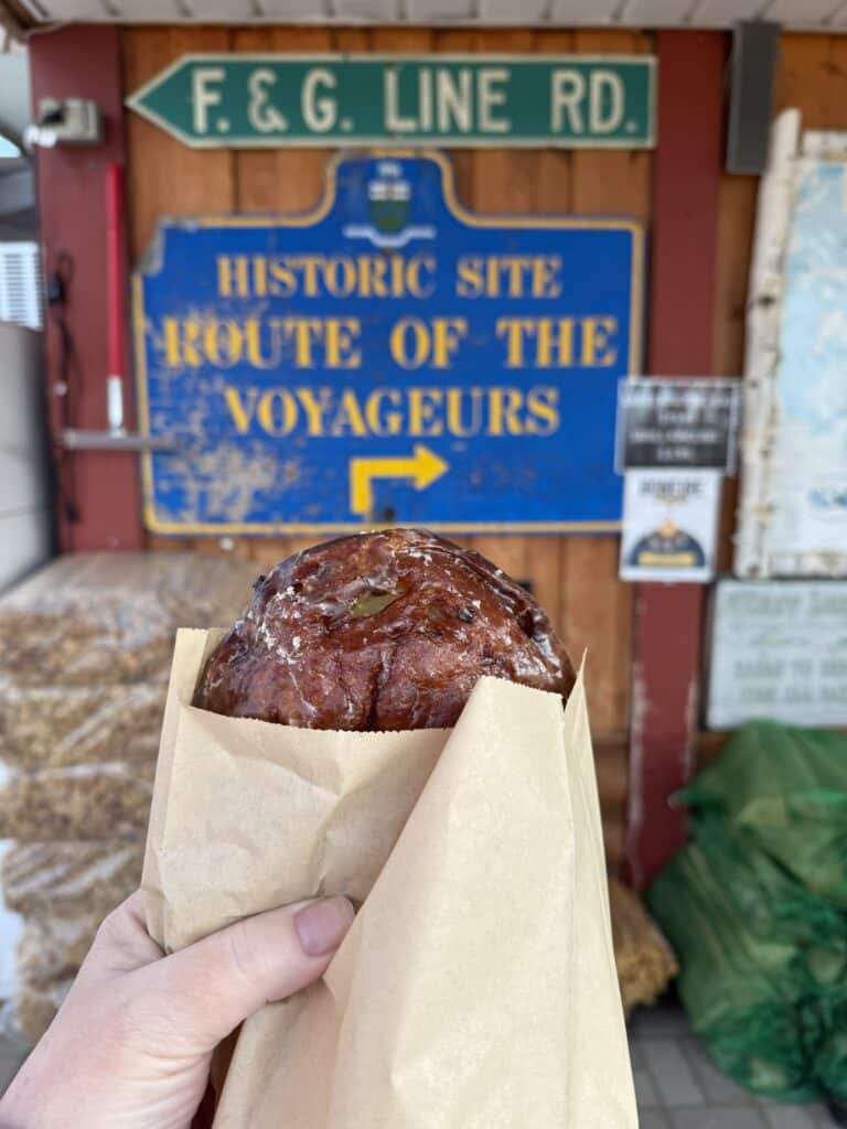 holding an apple fritter in a paper wrapper with a background of a blue sign with yellow lettering that reads "Historic Site Route of the Voyageurs"