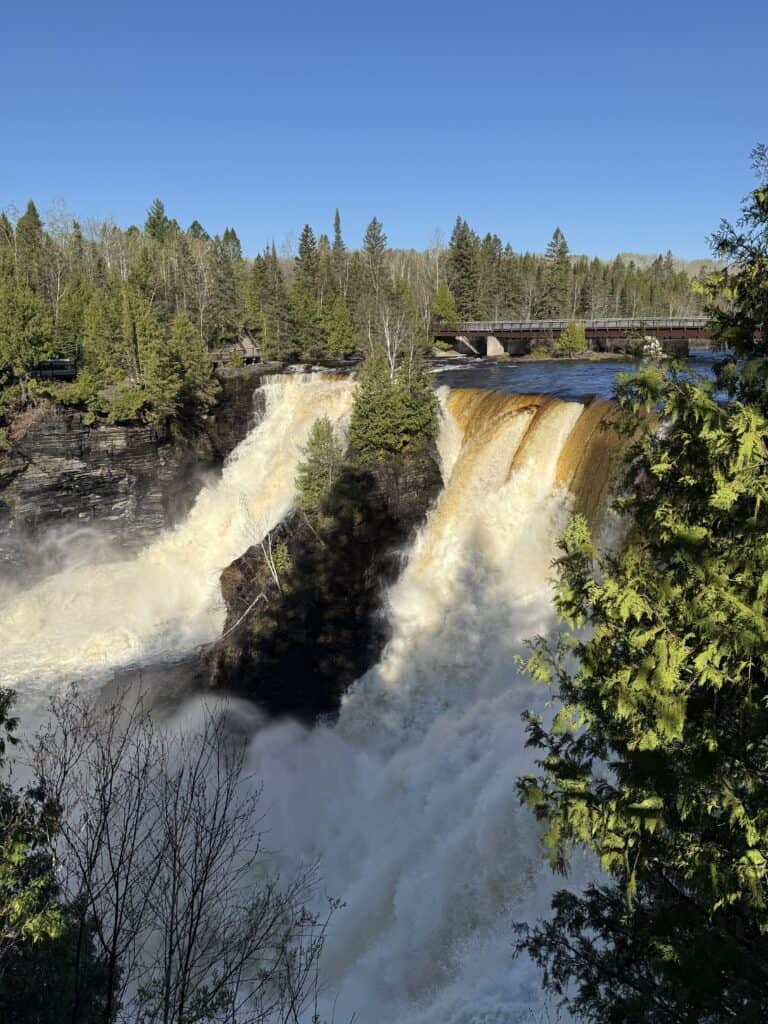 thundering waterfalls surrounded by evergreen trees with pedestrian bridge in background and bright blue sky.