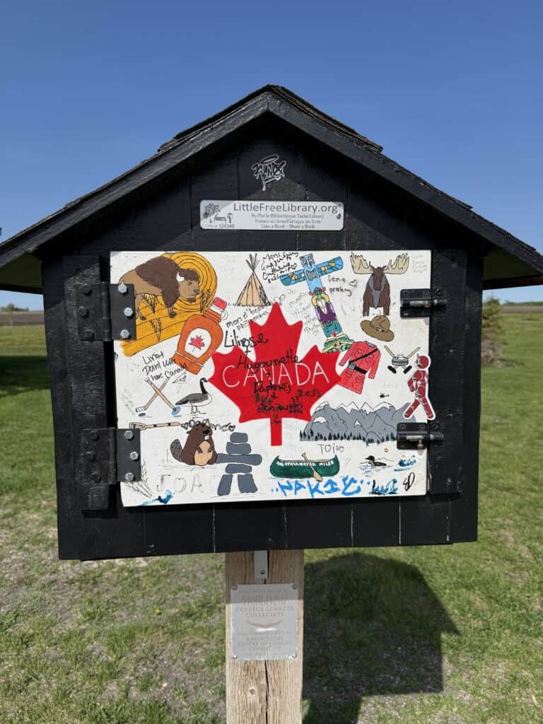 black wooden little free library with door painted with colourful images of Canada at the Centre of Canada Park in Manitoba.