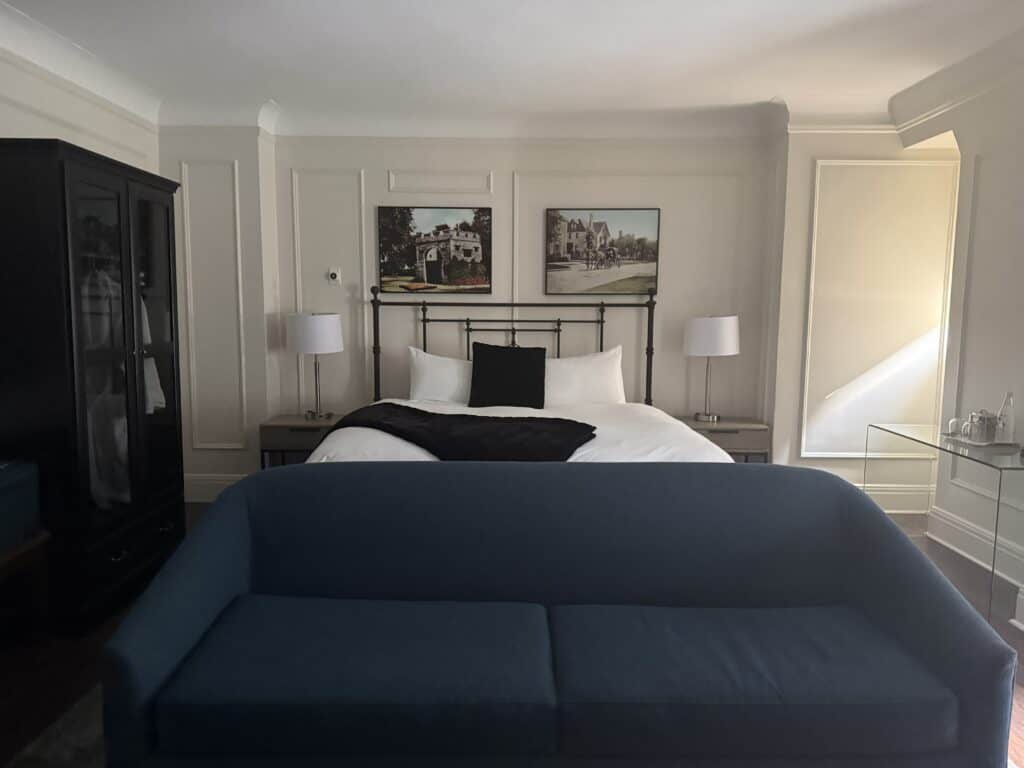 junior suite at the fort garry - white walls with panels - navy sofa in foreground and bed in background with black metal frame, white bedding with navy throw and pillow, two framed photos above bed and two side tables with white shaded lamps, dark armoir on left.