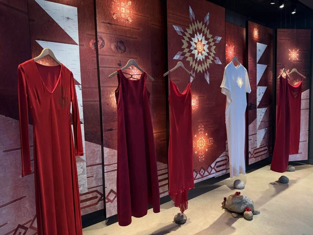 display of five red dresses and a white dress in the middle in front of wall with Indigenous symbols at the Canadian Museum for Human Rights.