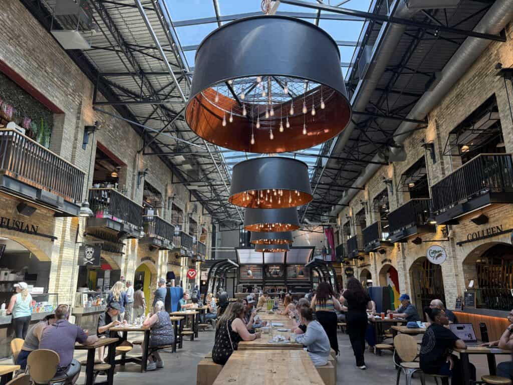 people eating at rows of tables in the forks market in winnipeg with large black chandeliers overhead