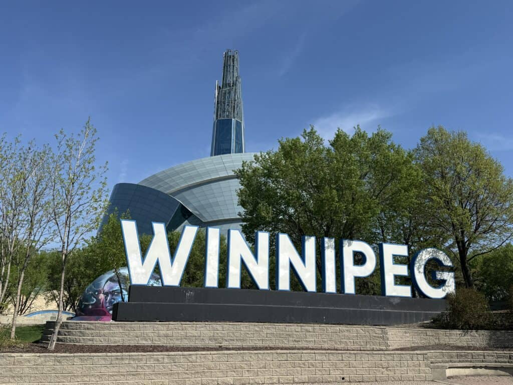 white "WINNIPEG" sign on platform with the Canadian Museum for Human Rights in the background and a bright blue sky.