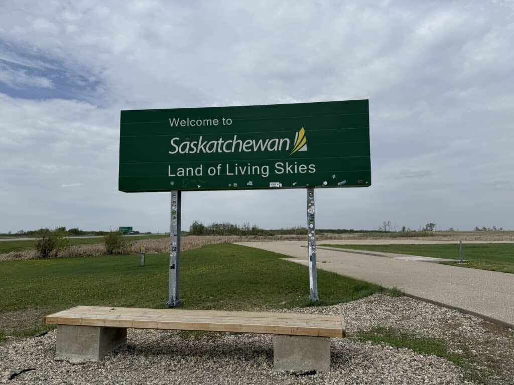 wooden bench in front of green highway sign reading "Welcome to Saskatchewan Land of Living Skies" on cloudy day.