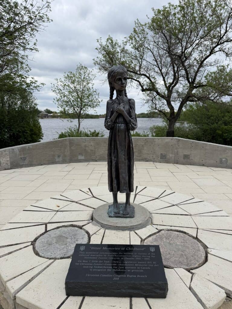 bronze statue of thin young girl in dress with hair braids and hands clasped over chest with lake and trees in background.