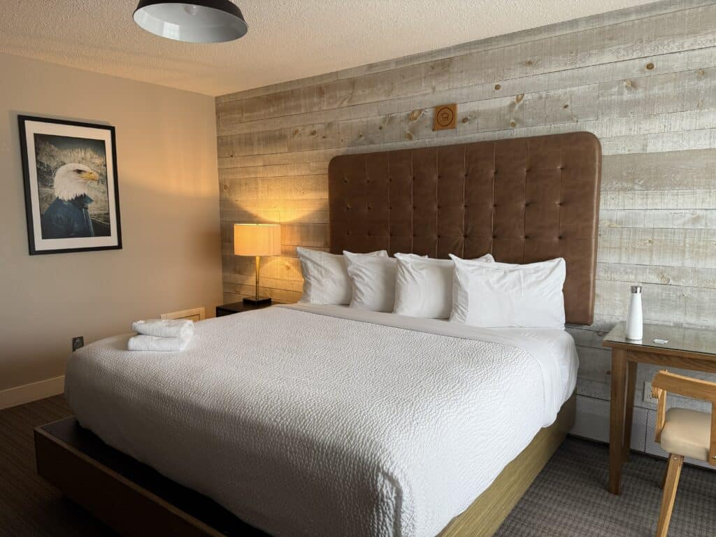 king size bed with white bedding and four pillows with brown upholstered headboard against barnboard wall.