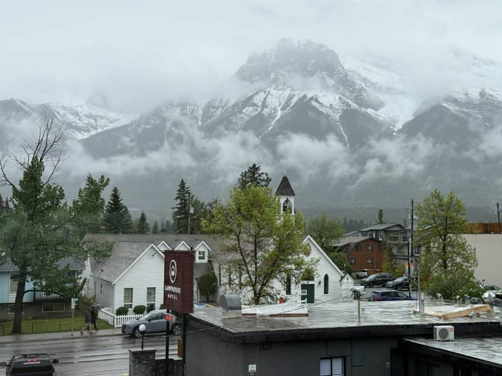 view of street in Canmore including white church with black steeple with low wispy white clouds and snow capped mountains in background