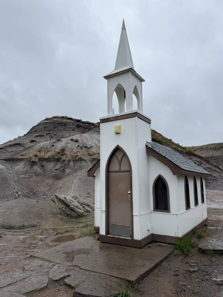 small white church with brown arched door, windows and tiles with rocky hill in background - Drumheller, Alberta