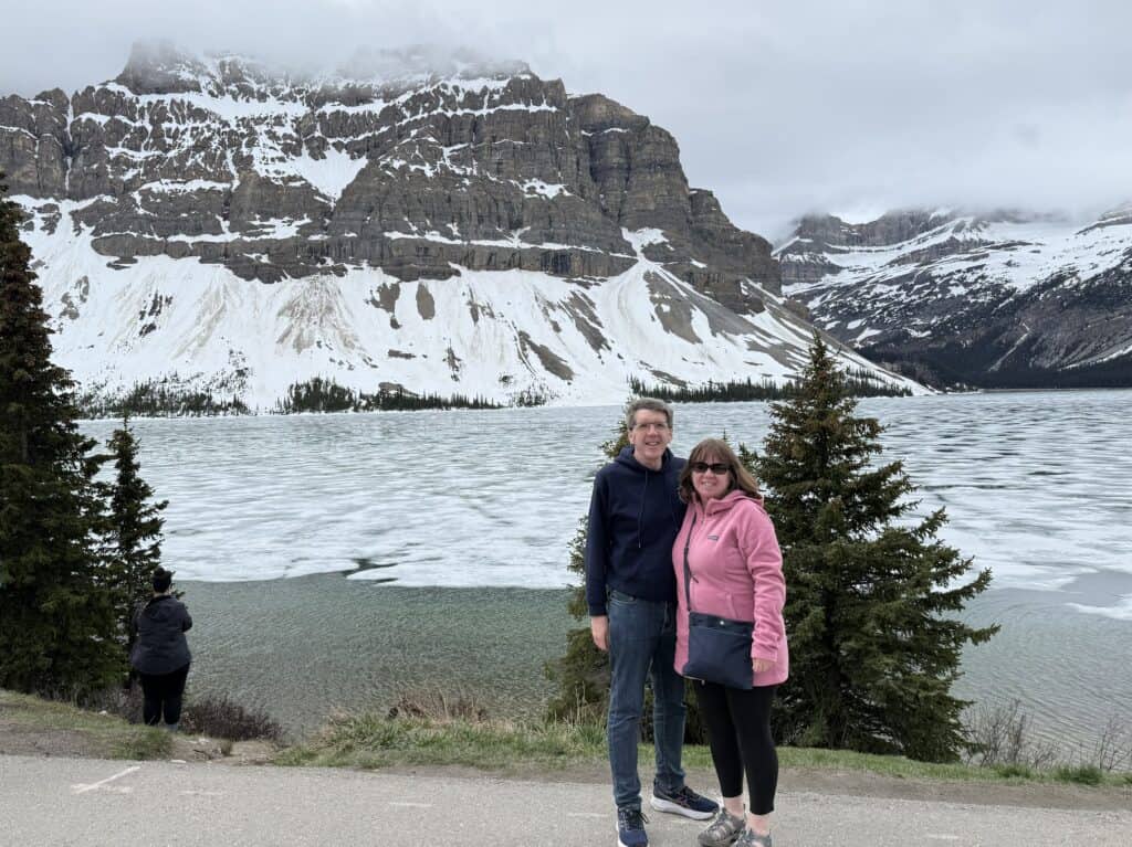 man in jeans and blue sweatshirt and woman in black pants, pink sweater and sunglasses posing at ice-covered Bow Lake with snow-covered mountains in background.