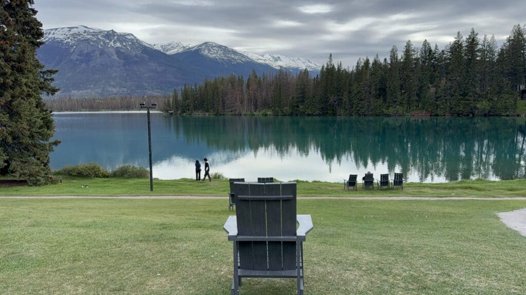 chairs on lawn at Jasper Park Lodge facing blue/green lake surround by evergreen trees and snow-capped mountains in background with overcast sky.