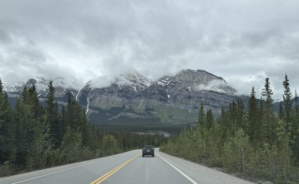 car on highway with evergreens on either side heading toward snow-capped mountains with low hanging clouds on an overcast day.