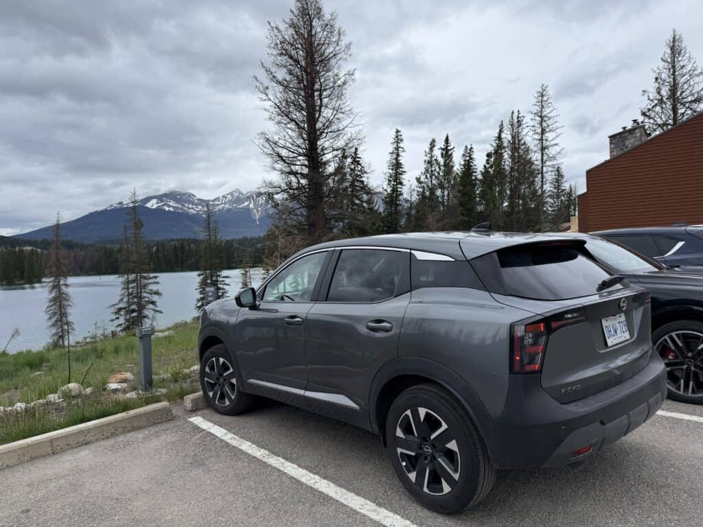 dark gray Nissan SUV parked in lot with evergreen trees, snow-capped mountains and lake in background at Jasper Park Lodge.