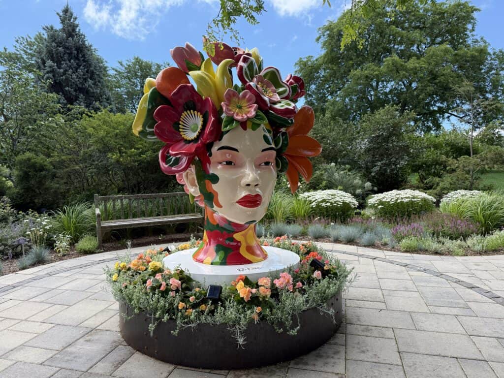 sculpture of woman's head with colourful red and flowers blooming on top set in gardens with shrubs, trees, flowers and a wooden bench in background.