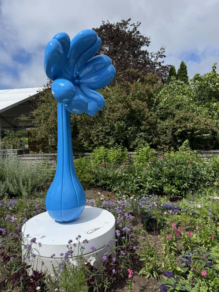 sculpture of single blue tulip on white pedestal set in garden with flowers and plants.