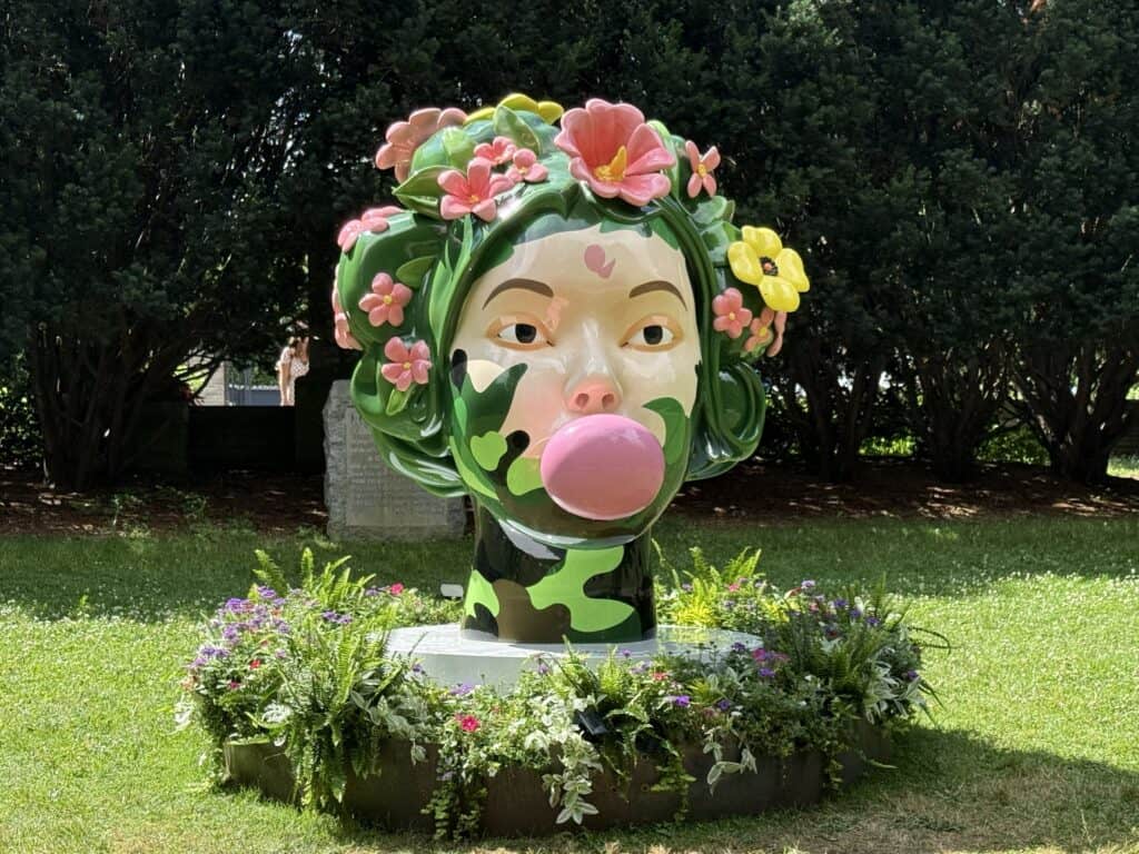 sculpture of female head with pink and yellow flowers and greenery on her head and blowing a large pink bubble.