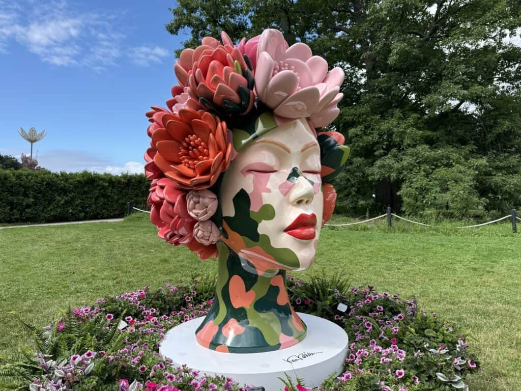 sculpture of female head with pink and coral flowers surrounded by flowers in garden