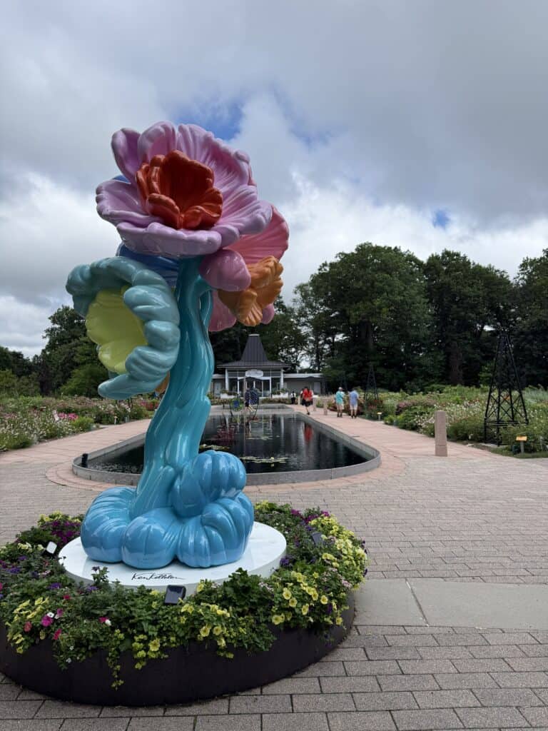 curvy flower sculpture with blue stem and colourful flowers in front of reflecting pond with teahouse in background at royal botanical gardens.