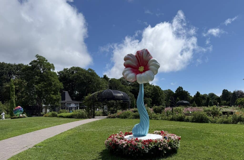 sculpture of single flower blossom with blue stem and pink and white petals surrounded by flower bed on lawn in hendrie park at the royal botanical gardens in burlington ontario.
