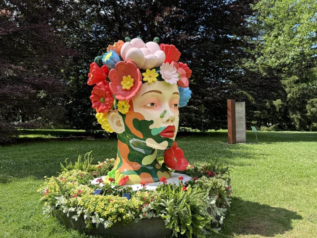 sculpture of woman's head with pink, red, yellow and blue flowers surrounded by flower bed on lawn with trees in background.