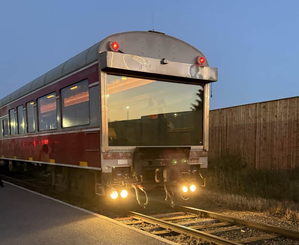 last car on train with large glass window with lights on parked on tracks at train depot at dawn.