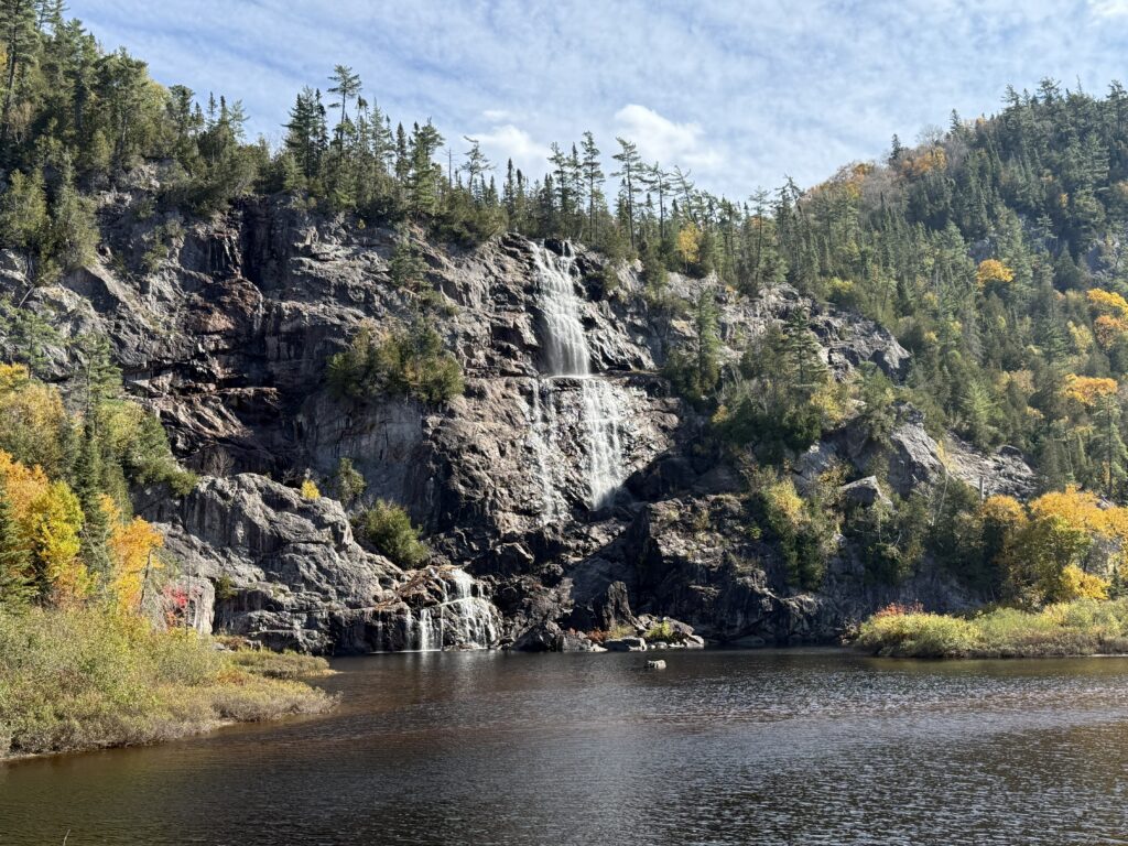waterfall over rocky cliff surrounded by evergreen trees and some trees with yellow leaves.
