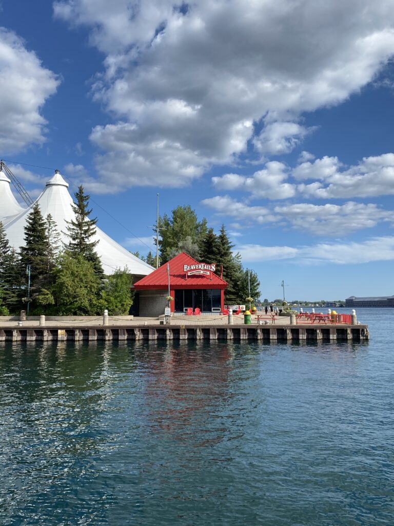 waterfront in sault ste. marie, ontario - red roofed BeaverTails outlet on pier with white tents of Roberta Bondar Park in background with bright blue sky and fluffy white clouds.